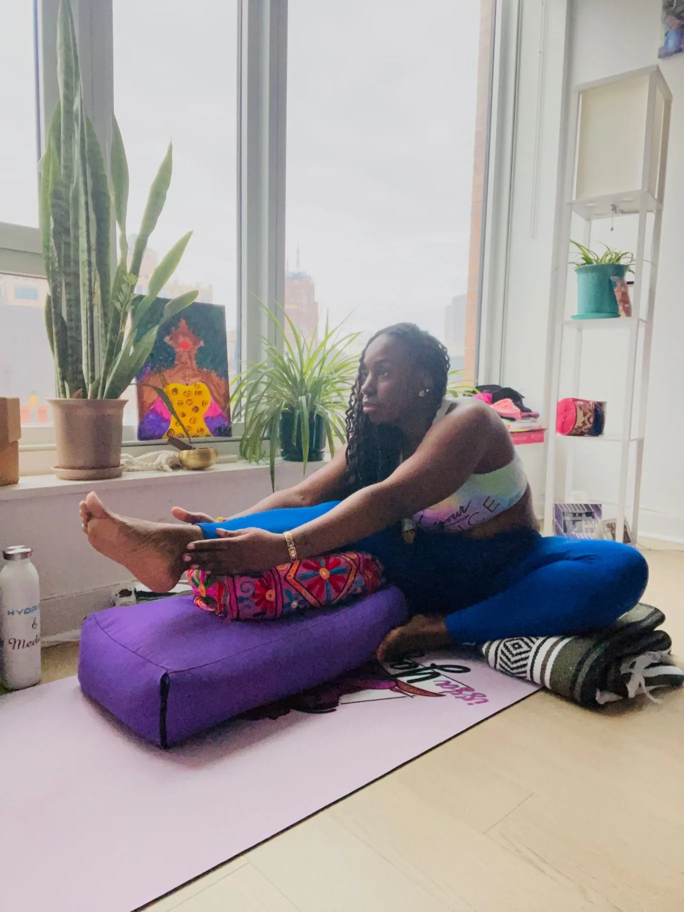 A woman practices a seated yoga stretch on a bolster in a bright, plant-filled room